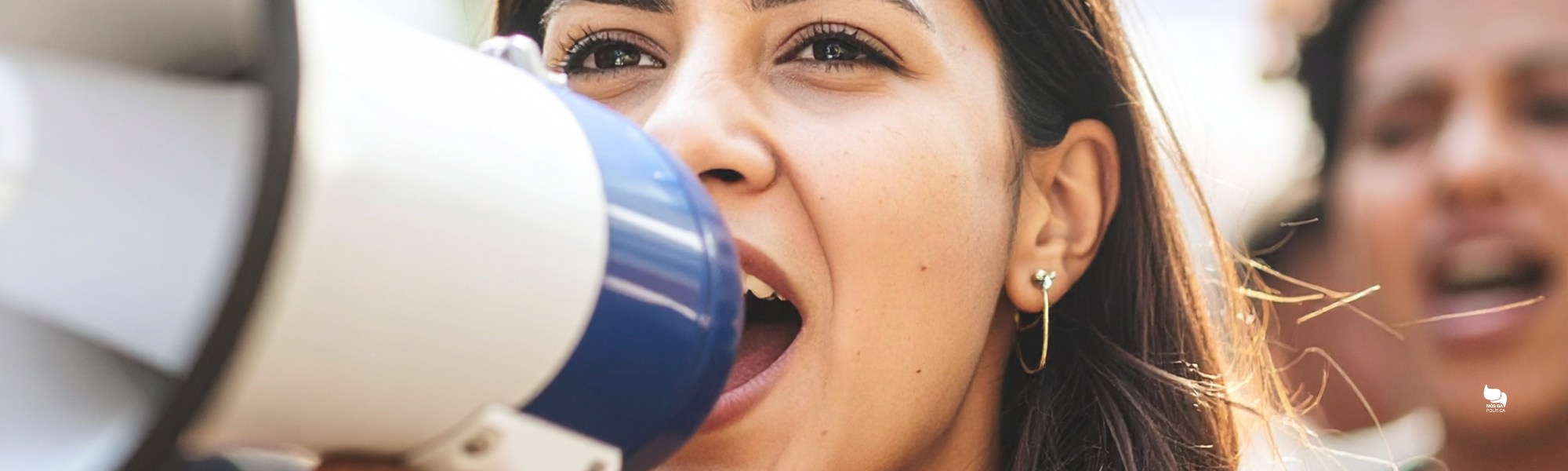 Mulher falando em um megafone durante manifestação pública, com expressão intensa, representando o uso do grito e da performance no discurso político.